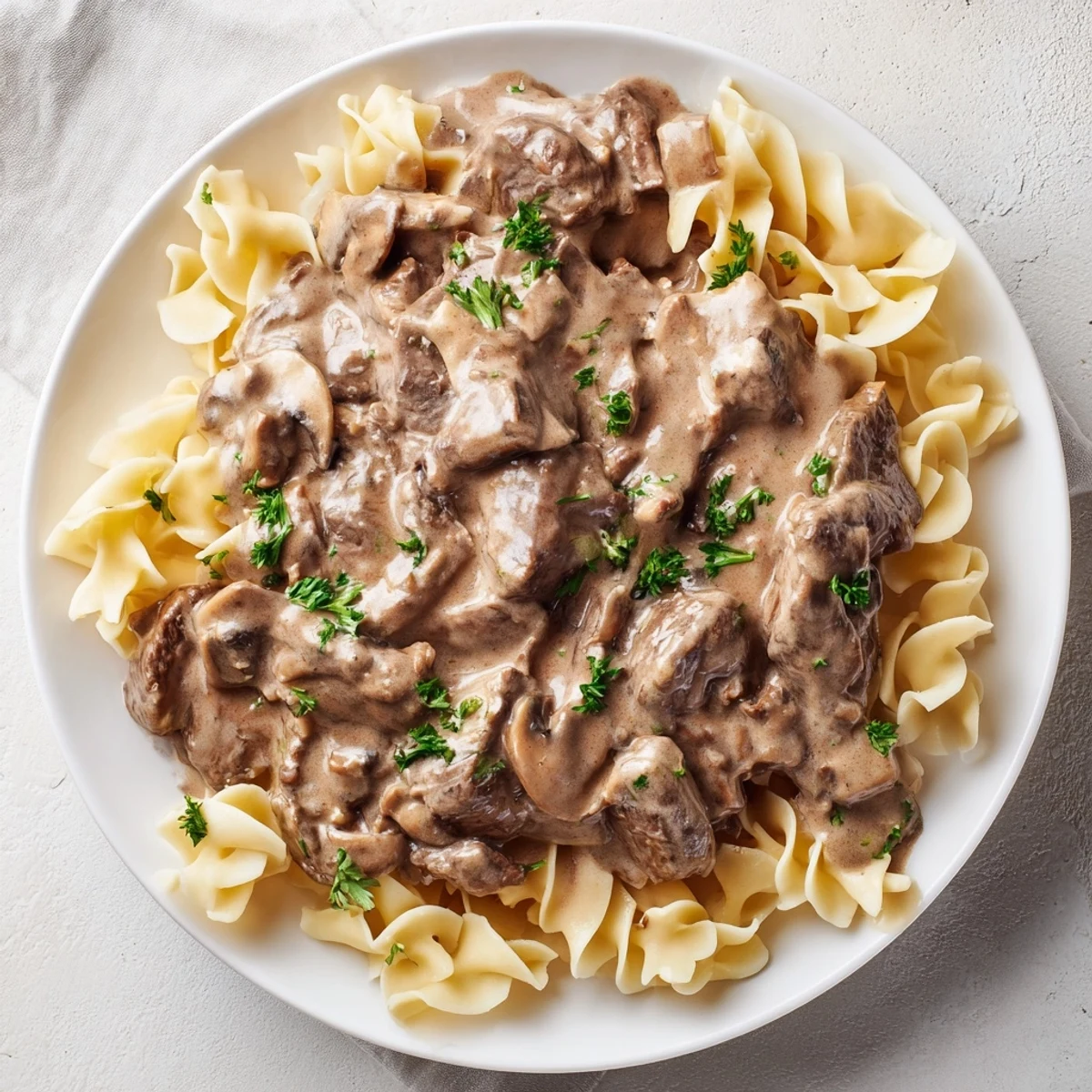 A close-up of creamy Beef Stroganoff with mushroom gravy ladled over golden egg noodles, garnished with fresh parsley.
