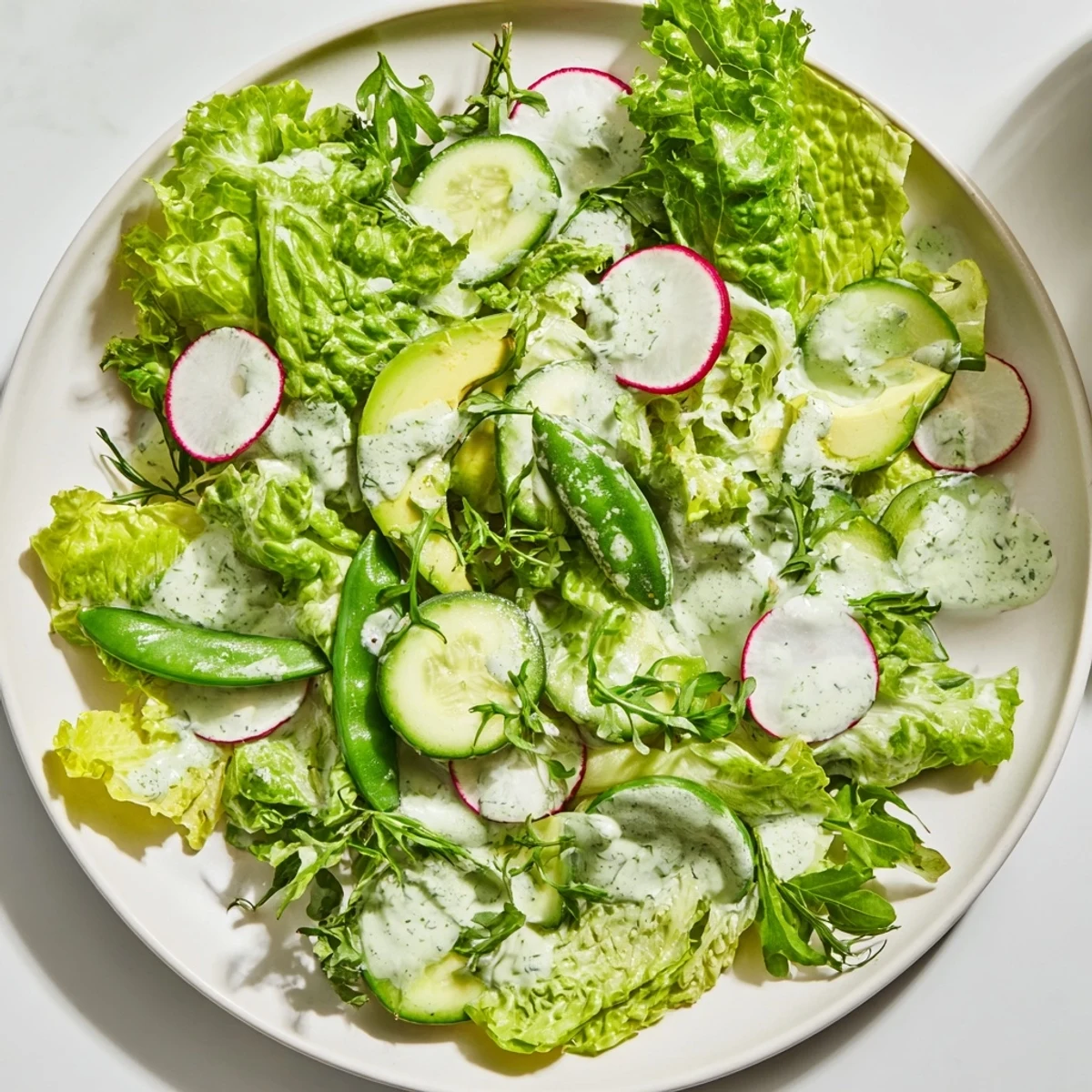 Green Salad with Green Goddess Dressing on a white plate, topped with sliced avocado, cucumber, radishes, and snap peas.