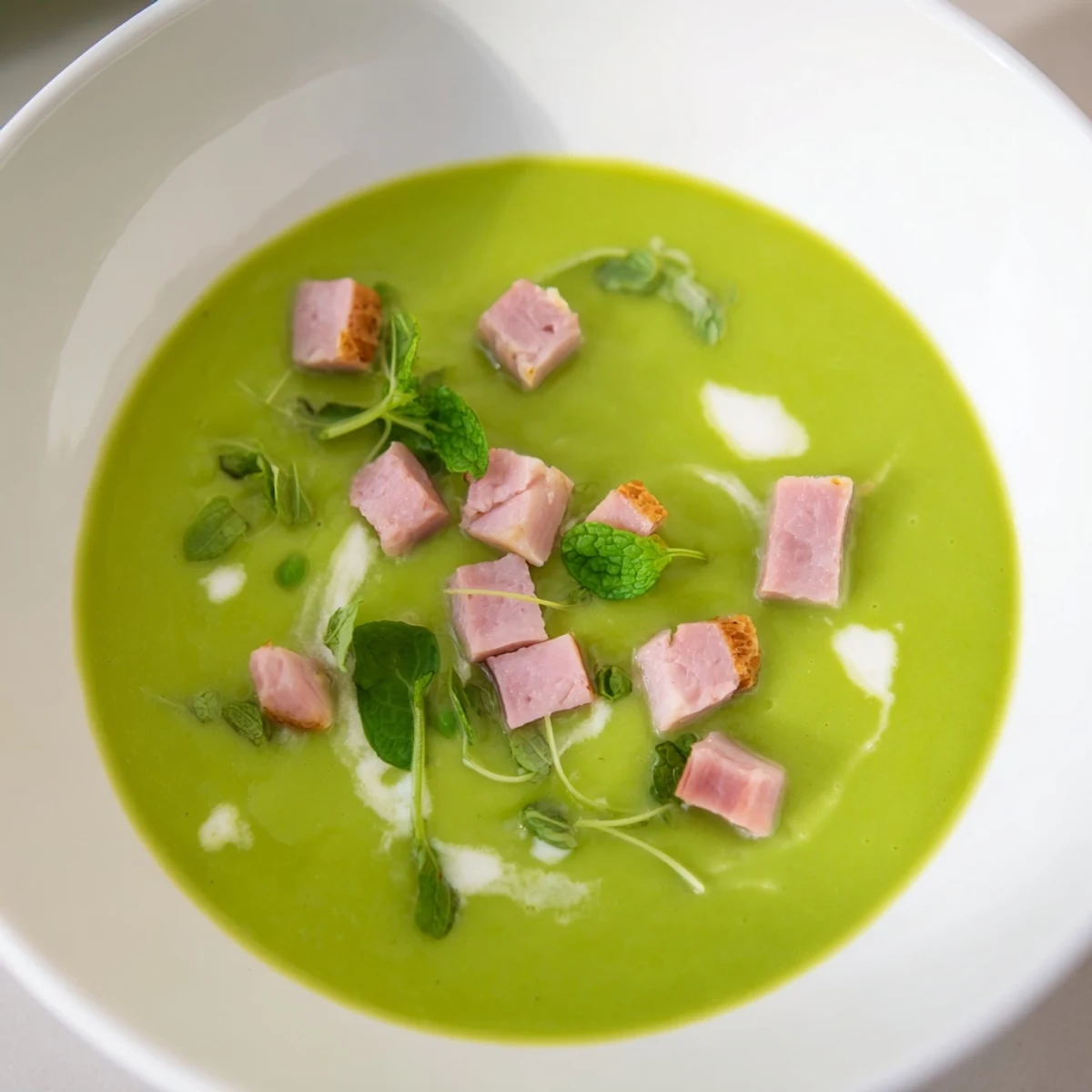 Freshly blended Spring Pea Soup with turkey ham garnished by pea shoots on a white surface.