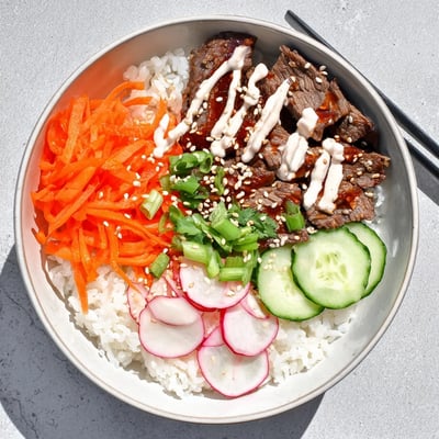 A close-up of Korean BBQ Steak Rice Bowls with Spicy Cream Sauce shows juicy steak slices, fresh carrots and cucumbers, and a rich sauce pooling on white rice.